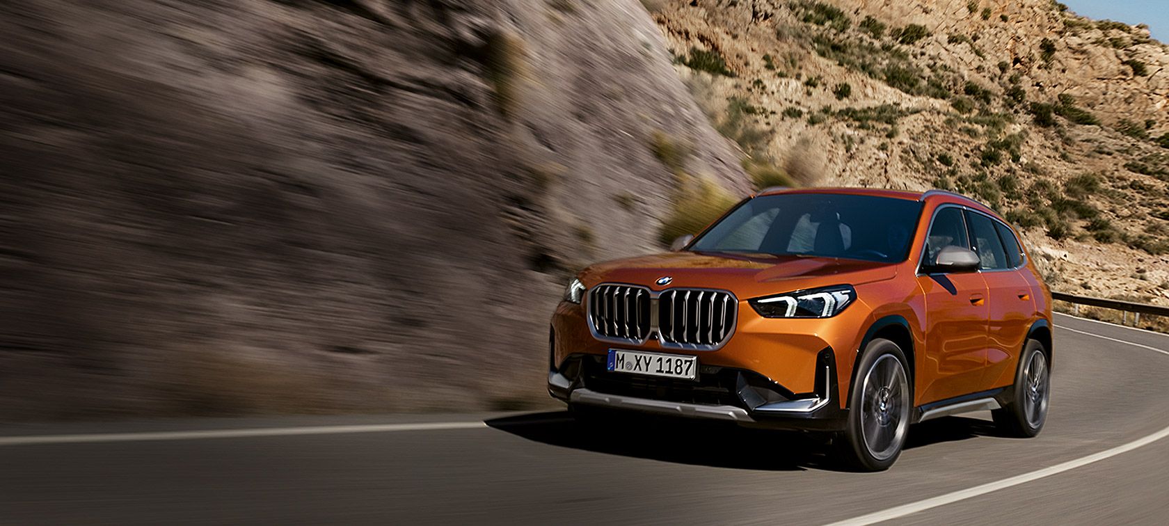 BMW X1 U11 xLine Utah orange metallic, vue de trois quarts avant sur route de campagne